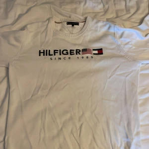 Vit långärmad tröja Tommy Hilfiger - Snygg vit långärmad tröja från Tommy Hilfiger med broderad logga och flaggor på bröstet. Rund hals med ribbad kant och diskreta detaljer i rött och blått. Knappt använd för har växt ifrån den väldigt snabbt. Perfekt för dig som gillar klassisk streetstyle och vill ha ett cleant plagg med tydlig branding. (Pris kan diskuteras)