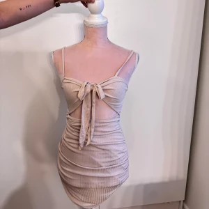 Beige ribbad kortklänning med knyt - Supersöt beige ribbad kortklänning med smala axelband och snygg knytning framtill. Klänningen har cut-out detalj under bysten och sitter tight. Perfekt för dig som gillar trendiga och feminina plagg med lite edge.
