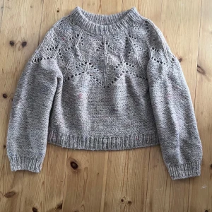 Jättefin hemstickad tröja - Supermysig beige stickad tröja med coolt hålmönster upptill och diskreta färgprickar i garnet. Tröjan har en bred ribbad kant vid hals, ärmslut och nederkant samt lång ärm och relaxed fit. Perfekt för lager på lager under kyliga dagar.