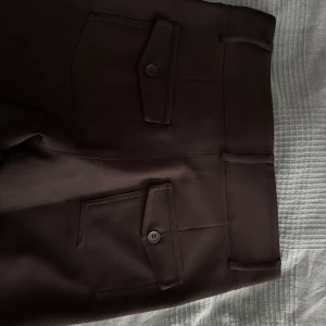 Svarta bootcut-kostymbyxor Gina Tricot S - Säljer ett par svarta bootcut-kostymbyxor från Gina Tricot i storlek S. Byxorna har snygga fickor med lock och knapp bak samt hällor för bälte. De är tillverkade i ett mjukt material med lite stretch och har en klassisk, stilren look. Har använt 2 gånger. Sitter tajt och formar kroppen. Pris kan diskuteras.