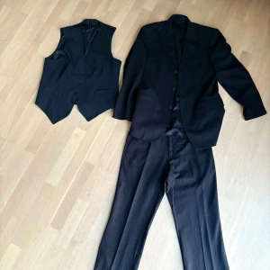 Svart kostym från Conwell, tre delar - Säljer en klassisk svart kostym från Conwell med kavaj, väst och byxor. Kostymen har diskreta ränder, treknäppt väst och raka byxor. Perfekt för dig som vill ha en stilren och tidlös look. Materialet känns mjukt och följsamt.