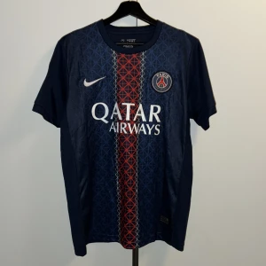 PSG Dembele #10 matchtröja Nike - Paris Saint-Germain fotbollströja med O. Dembélé och nummer 10 på ryggen. Marinblå med röd och vit randig detalj i mitten, PSG-logga och Qatar Airways tryck på bröstet. Tillverkad i lätt och ventilerande polyester, perfekt för träning eller match.