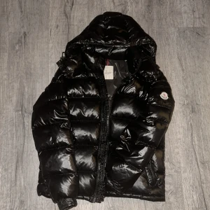 Moncer maya svart - Storlek 1- Säljer en svart, glansig pufferjacka från Moncler med huva och dragkedja. Jackan har det klassiska Moncler-märket på ärmen och coola detaljer som quiltad design och innerfoder med serietidningsmönster. Perfekt för kalla dagar när du vill hålla stilen.