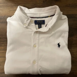 Vit skjorta från Polo Ralph Lauren - Vit skjorta från Polo Ralph Lauren med klassisk krage, knappar framtill och broderad marinblå logga på bröstet. På manschetten finns en snygg broderad text. Skjortan är i bomull och har en stilren, tidlös look som passar perfekt till jeans eller chinos.