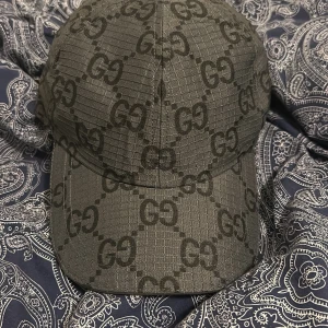Svart keps från Gucci med GG-mönster - Snygg svart keps från Gucci i storlek M, tillverkad i Italien. Kepsen har det klassiska GG-mönstret över hela ytan och är gjord i ett slitstarkt syntetmaterial. Justerbar baktill och med diskreta sömmar för en clean look. Perfekt för dig som gillar exklusiva accessoarer.
