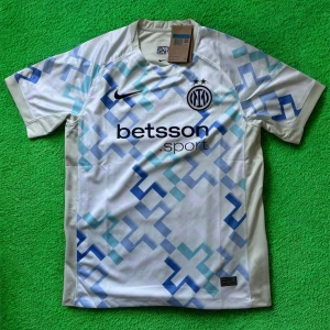 Inter Milan vit/blå Nike fotbollströja - Snygg Inter Milan fotbollströja från Nike i vit med ljusblå och marinblå geometriskt mönster. Kortärmad modell med rund hals och tryckt sponsorlogga på bröstet. Materialet är lätt och andas, perfekt för träning eller match. Klubbmärket och Nike-loggan är broderade.