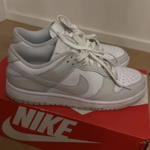 Nike Dunk Low vit/grå sneakers - Säljer ett par fräscha Nike Dunk Low sneakers i vitt och ljusgrått skinn. Klassisk siluett med perforerad tå, snörning och låg profil. Perfekta för dig som gillar stilrena och cleana sneakers med ikonisk Nike Swoosh på sidan.