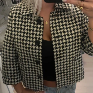 Rutig kavajjacka i svart och vitt - Snygg kavajjacka med klassiskt hundtandsmönster i svart och vitt. Jackan har krage, stora svarta knappar framtill och trekvartsärmar. Modellen är boxig och croppad, perfekt att slänga över en enkel topp för en cool look.