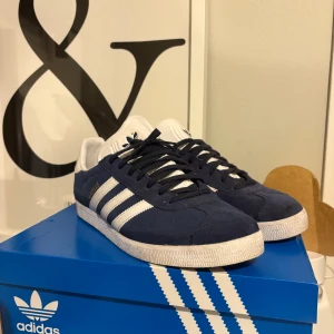 Adidas Gazelle blå mockasneakers - Snygga Adidas Gazelle sneakers i marinblå mocka med vita detaljer och klassiska tre ränder på sidorna. Skorna har vit sula, snörning och guldtexten 'GAZELLE' på sidan. Perfekta för dig som gillar retrostil och streetwear. Kommer i original boxen! Nypris:1349kr Mitt pris: 799