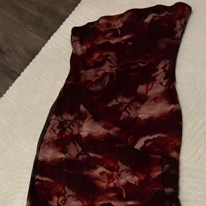 Vinröd omlott långklänning - Säljer en vinröd långklänning med blommigt mönster och omlott-detalj. Klänningen är axelbandslös och har en passform som framhäver figuren. Materialet är lätt och skir polyester, perfekt för dig som vill sticka ut med en elegant look.