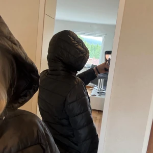 Svart pufferjacka med huva - Säljer en svart pufferjacka med huva och dragkedja framtill. Jackan har quiltad design med horisontella sömmar och två sidofickor med dragkedja. Perfekt för kyliga dagar och enkel att matcha med jeans eller mjukisbyxor. Jackan har små hål vid armhålan men inget som man märker