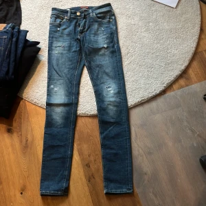 Blå skinny jeans med slitningar - Säljer ett par blå jeans från Jack and Jones i modellen Liam och i storleken 29 32. Jeansen har snygga slitningar och lätt tvättade detaljer. Jeansen har klassisk femficksdesign, normal midja och smal passform hela vägen ner. Perfekta för dig som gillar en trendig och avslappnad look.