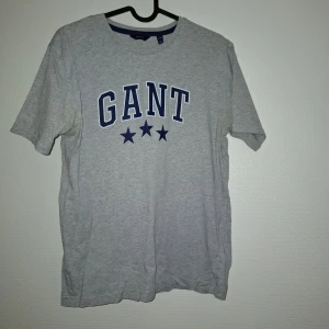 Grå GANT t-shirt med blå logga - Snygg grå t-shirt från GANT med klassisk blå logga och tre stjärnor framtill. T-shirten har rund halsringning med blå kant och är kortärmad. Perfekt för en clean och sportig look. Tillverkad i mjuk bomull för skön komfort.