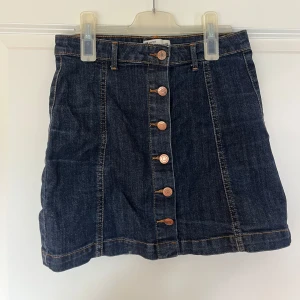 Mörkblå jeanskjol med knappar - Snygg mörkblå jeanskjol med raka linjer och knappar hela vägen framtill. Kjolen har bälteshällor och klassiska sömmar i kontrastfärg. Från Gina Tricot, aldrig använd