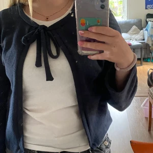 Mörkblå kofta med knyt framtill - Säljer en mörkblå kofta med knytband vid halsen. Koftan har långärmade ärmar och är gjord i mjukt material som känns skönt mot huden. Perfekt att slänga över en t-shirt för en chill och enkel look.