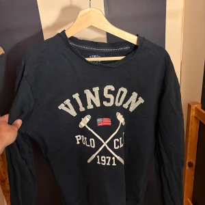 Mörkblå tröja Vinson Polo Club - Mörkblå långärmad tröja från Vinson Polo Club med tryck framtill, amerikansk flagga och korsade poloklubbor. Rund halsringning och mjukt bomullsmaterial. Perfekt för dig som gillar sportig och avslappnad stil.