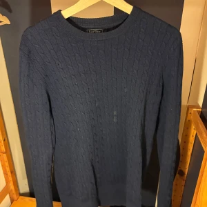 Mörkblå kabelstickad tröja East West - Kabelstickad mörkblå tröja från East West i lammull. Tröjan har rund hals, långa ärmar och snygga bruna mockadetaljer på armbågarna. Perfekt för dig som gillar klassisk stil med en twist.