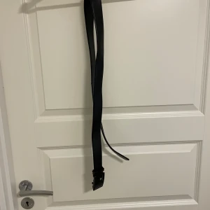 Svart bälte från Dongfang BAOLUO - Stilrent svart bälte från Dongfang BAOLUO med en rektangulär, blank spänne i svart med logga och text. Bältet är slätt och har en klassisk look som passar till många outfits. Perfekt accessoar för att lyfta din stil.