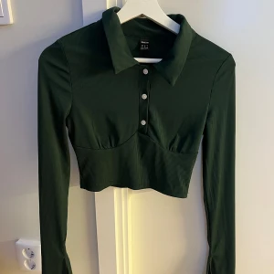 Mörkgrön croppad polotopp från Shein - Säljer en mörkgrön croppad polotopp från Shein med långa ärmar, krage och tre vita knappar framtill. Toppen är ribbad och har en figurnära passform som framhäver midjan. Perfekt för dig som gillar trendiga och stilrena plagg.