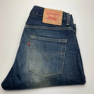 Levi's 507 blå jeans - Klassiska blå Levi's 507 jeans med snyggt slitna detaljer och ikonisk läderpatch bak. Jeansen har raka ben och normal passform, tillverkade i robust denim med kontrastsömmar och femficksdesign. Perfekta för en avslappnad och tidlös stil.