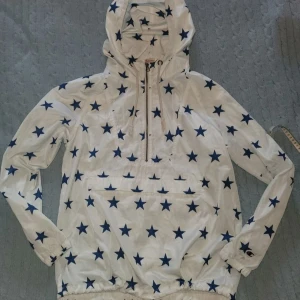Champion vindjacka med blå stjärnor XL - Snygg vit vindjacka från Champion med blå stjärnmönster över hela jackan och stort Champion-logga på ryggen. Jackan har huva, half zip framtill och praktisk ficka. Tillverkad i lätt polyester, perfekt för dig som gillar streetwear och sportig stil.