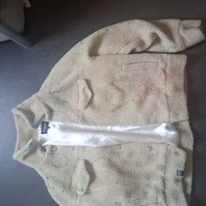 Beige teddyjacka från 5ive6ix - Mysig beige teddyjacka från 5ive6ix med två bröstfickor och knappar framtill. Jackan har en fluffig struktur och är fodrad med glansigt vitt tyg. Perfekt för dig som gillar en chill och trendig look.