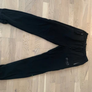 Svarta mjukisbyxor från BOSS - Svarta mjukisbyxor från BOSS med dragsko i midjan och muddar vid bensluten. Byxorna har fickor med dragkedja och en diskret BOSS-logga på låret. Perfekta för chill eller träning.