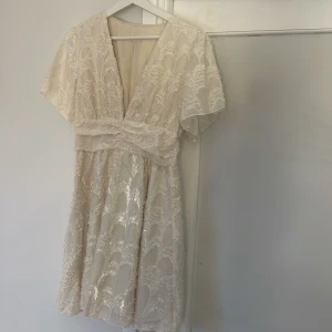 Champagnefärgad kortklänning med spets - Superfin kortklänning i champagnefärg med v-ringning och voluminösa korta ärmar. Klänningen har ett vackert spetsmönster och markerad midja med resår. Materialet känns lätt och luftigt, perfekt för sommarens alla tillfällen. Oandvänd så nyskick.