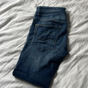 Blå jeans från Jack and jones  - Snygga blå jeans från Jack and jones med klassisk femficksdesign och kontrastsömmar. Jeansen har en rak passform och är tillverkade i slitstarkt denim av bomull. Perfekta för dig som gillar en tidlös och stilren look.