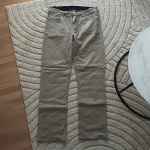 Beige chinos från Cotton Team W34 L32 - Snygga beige chinos från Cotton Team med klassisk femficksdesign och raka ben. Byxorna är tillverkade i bomull med lite stretch för extra komfort. Perfekta för en clean och avslappnad stil. Passar dig som gillar stilrena och enkla byxor.