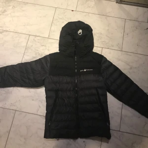 Svart dunjacka från Sail Racing M - Säljer en svart dunjacka från Sail Racing i storlek medium. Jackan har huva, dragkedja framtill och två sidofickor med dragkedja. Fylld med 800 fill power dun från Allied Feather + Down. Perfekt för kalla dagar och riktigt snygg streetstil.