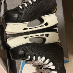 Svarta Bauer skridskor Supreme - Säljer ett par svarta Bauer Supreme skridskor med vita snören och detaljer i gult. Ovandelen är i syntetmaterial och skinn, med robust konstruktion och klassisk design. Perfekta för dig som vill ha snygga och bekväma skridskor till isen.