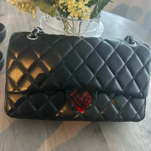 Svart quiltad axelväska med kedja - Snygg svart axelväska i quiltad design med silverfärgad kedjerem. Väskan är tillverkad i skinn och har ett klassiskt rutmönster. Perfekt storlek för det viktigaste och ger en stilren vibe till din outfit.