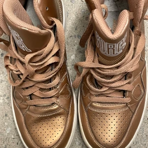 Bronsfärgade Nike Air Force sneakers - Nike Air Force sneakers i metallic brons med högt skaft och klassisk snörning. Skorna har perforerad tå för extra ventilation och vit sula som kontrast. Snyggt FORCE-tryck på plösen och cool retrovibe. Perfekta för dig som vill sticka ut med din stil.