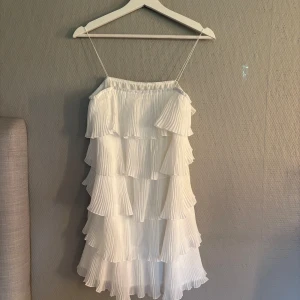 Vit volangklänning med tunna axelband - Supersöt vit klänning med flera lager volanger och tunna axelband. Plisserat tyg ger en luftig och flowy känsla, perfekt för varma dagar. Klänningen är kort och har en romantisk vibe som passar dig som gillar en feminin stil.