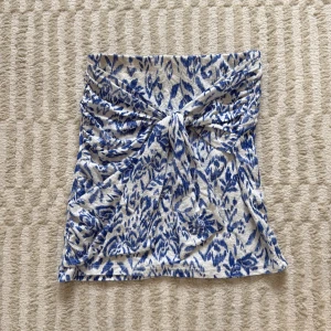 Strapless topp med knytdetalj från Gina Tricot - Supersöt topp från Gina Tricot i vitt och blått med blommigt mönster. Kjolen har en snygg knyt-detalj framtill och är gjord i ett mjukt, stretchigt material. Perfekt för dig som vill ha en trendig och bekväm look.