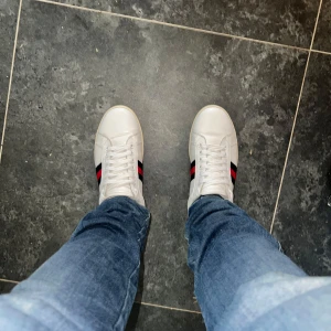Gucci ace  - Säljer ett par klassiska vita sneakers från Gucci i skinn med den ikoniska röd- och blåa sidoremsan. Skorna har rund tå, platt sula och snörning framtill. Perfekta för dig som vill ha en clean och lyxig look med streetkänsla.