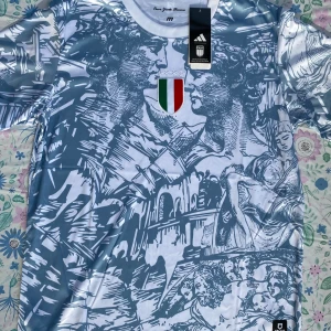 Italien Adidas fotbollströja M - Snygg kortärmad Italien fotbollströja från Adidas med konstnärligt tryck i blått och vitt, samt broderat italienskt klubbmärke med stjärnor på bröstet. Materialet är lätt och andas, perfekt för match eller träning. Rund hals och officiell licens.