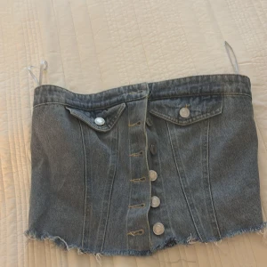 Jeans tubtopp med knappar och fickor - Säljer en cool jeans tubtopp med silvriga knappar framtill och två fickor med lock. Toppen har rå kant nedtill och är axelbandslös med en bandeauform. Perfekt för dig som gillar denim och vill sticka ut med en edgy look.