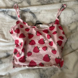 Rosa jordgubbsmönstrad linnetopp Shein - Söt rosa linnetopp från Shein med jordgubbsmönster över hela tyget. Toppen har smala axelband och croppad passform med vågig kant nedtill. Perfekt för dig som gillar färgglada och lekfulla plagg. Materialet är mjuk bomull.