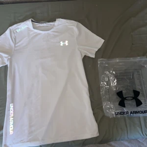 Vit Under Armour t-shirt  - Snygg vit t-shirt från Under Armour med reflekterande logga på bröstet och texten UA-SPEC samt UNDER ARMOUR i holografisk stil. Modellen är kortärmad och har rund hals. Perfekt för löpning och annan träning, tillverkad i lätt polyester som andas.