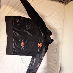 Svart Adidas Chile 62 jacket - Säljer en svart Adidas vindjacka/kofta med glansig finish och färgglada detaljer. Jackan har dragkedja, långärmade ärmar och broderad Adidas-logga i orange, blått och vitt på ryggen. Snygga kontrastlinjer längs ärmarna och praktisk ficka med dragkedja framtill. Pris går att diskutera! Hoppas du vill köpa :)