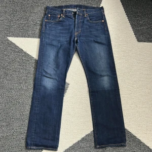 Levi's 501 blå jeans W30 L30 - Klassiska Levi's 501 jeans i mörkblå denim med raka ben och fem fickor. Snygga kontrastsömmar och ikonisk läderpatch bak i midjan. Passar dig som gillar tidlös stil och vill ha ett par jeans som funkar till allt. Modellen har normal passform och knappgylf.