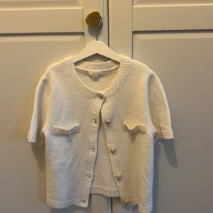 Vit kortärmad kofta från Gina Tricot - Supersöt vit kofta från Gina Tricot med korta ärmar och snygga guldfärgade knappar framtill och på de två dekorativa fickorna. Ribbstickade detaljer vid hals, ärmslut och nederkant ger en chill vibe. Perfekt att slänga över en topp för en clean look. Hör av er för fler bilder.