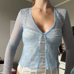 Ljusblå spetsblus med knappar - Säljer en ljusblå blus i transparent spets med blommigt mönster. Blusen har lång ärm, v-ringad hals och knappar framtill. Den är croppad och har fina vågiga kanter nedtill. Perfekt för dig som gillar romantiska detaljer och vill sticka ut.