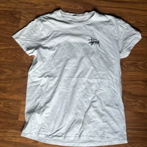 Vit Stüssy t-shirt med tärningar - Vit t-shirt från Stüssy med svart logga på bröstet och stort tryck med tärningar på ryggen. Klassisk rund hals och korta ärmar. Skön streetwear-vibe och tillverkad i mjuk bomull. Perfekt för dig som gillar clean stil med coola detaljer.