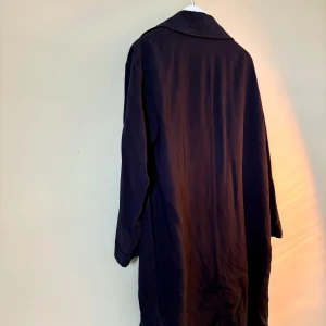 Svart lång trenchcoat från Zara - Snygg svart trenchcoat från Zara med klassisk krage och långa ärmar. Jackan har en loose passform och är tillverkad i ett mjukt bomullsmaterial. Perfekt för dig som gillar stilrena och tidlösa plagg med lite extra längd.