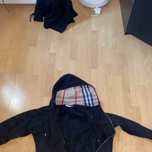 Svart Burberry vindjacka XXL - Svart vindjacka från Burberry i storlek XXL. Jackan har huva med klassiskt Burberry-rutigt foder, guldfärgad dragkedja med Burberry-logga och snörning vid huvan. Perfekt för dig som vill ha en stilren jacka med lyxiga detaljer och ikoniskt mönster.