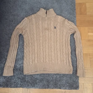 Beige stickad polotröja Polo Ralph Lauren - Säljer en beige stickad polotröja från Polo Ralph Lauren med klassiskt kabelstickat mönster och half zip i halsen. Tröjan har långärmad passform, ribbade muddar och den ikoniska Polo-loggan broderad på bröstet. Perfekt för dig som gillar stilren och tidlös design.