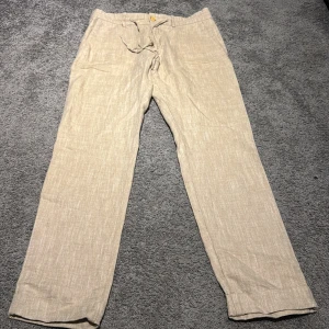 Beige linnebyxor från Morris 29/32 - Snygga beige linnebyxor från Morris i rak modell med normal passform. Byxorna har knytband i midjan och en klassisk look som funkar till chillare dagar. Materialet är linne vilket gör dem luftiga och sköna. Perfekt för dig som gillar stilren och enkel design.
